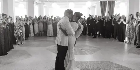 A black and white photograph of a bride and groom sharing a first dance on a reception floor while surrounded by guests.