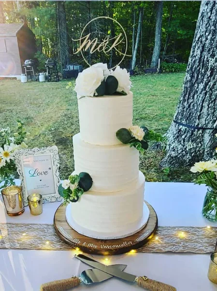 A three-tiered white wedding cake decorated with white roses and greenery, topped with a gold monogram, sitting on a wooden stand.