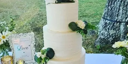 A three-tiered white wedding cake decorated with white roses and greenery, topped with a gold monogram, sitting on a wooden stand.