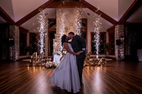 Bride and groom dancing in a wood-beamed indoor wedding reception hall with sparkler fountains and illuminated Mr & Mrs letters.