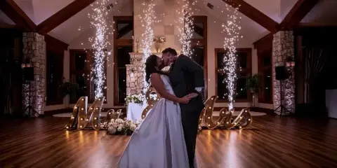 Bride and groom dancing in a wood-beamed indoor wedding reception hall with sparkler fountains and illuminated Mr & Mrs letters.