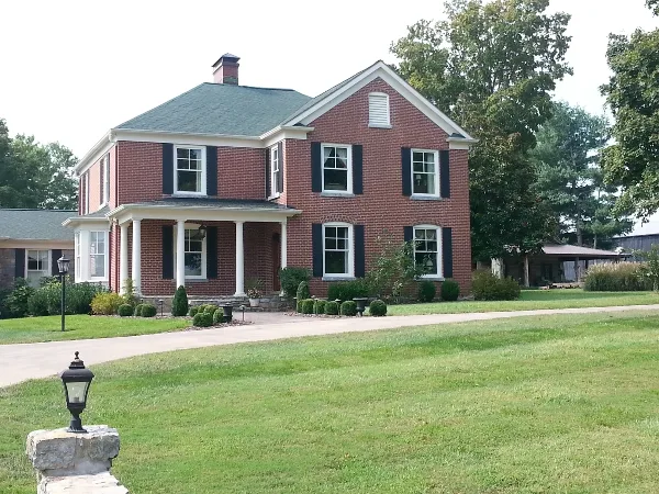 Exterior view of a two-story red brick historic house with white trim and a columned porch, set on a large green lawn.