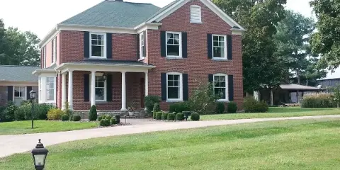 Exterior view of a two-story red brick historic house with white trim and a columned porch, set on a large green lawn.