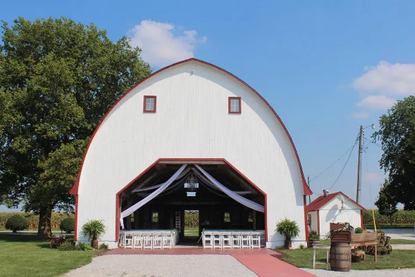 Exterior view of a white and red arched wedding barn with white chairs and drapery set up inside the open entrance.