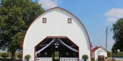 Exterior view of a white and red arched wedding barn with white chairs and drapery set up inside the open entrance.