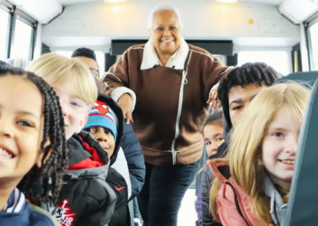 A smiling bus driver standing in the aisle of a bus surrounded by passengers.