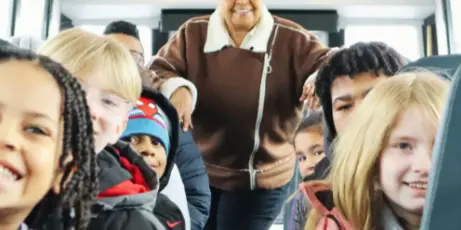 A smiling bus driver standing in the aisle of a bus surrounded by passengers.