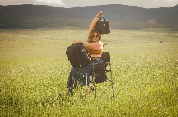A woman sitting in a makeup chair in a grassy field with mountains in the background, holding up a makeup bag.