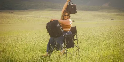 A woman sitting in a makeup chair in a grassy field with mountains in the background, holding up a makeup bag.