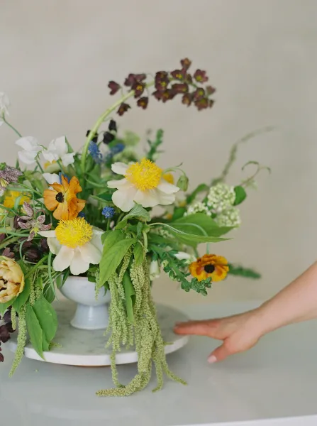 A hand adjusting a vibrant floral arrangement featuring white and yellow peonies, orange ranunculus, and greenery in a white vase.