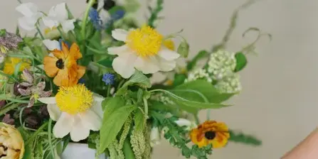 A hand adjusting a vibrant floral arrangement featuring white and yellow peonies, orange ranunculus, and greenery in a white vase.