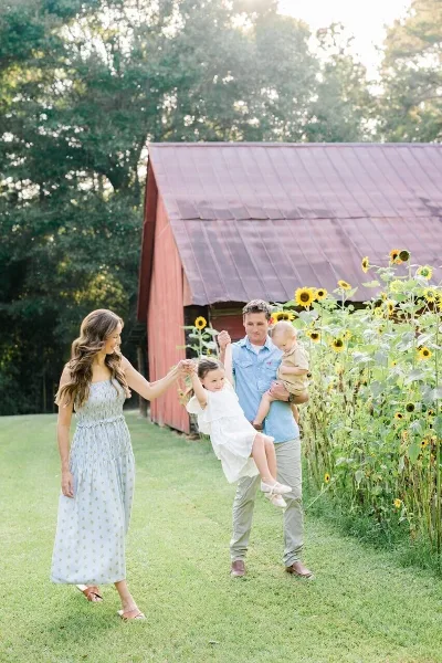 A family of four poses outdoors near a red barn and sunflowers, with the father swinging his daughter.
