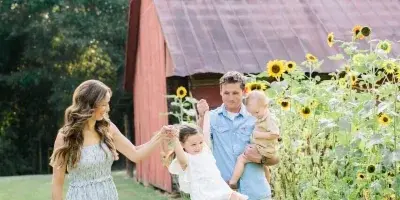 A family of four poses outdoors near a red barn and sunflowers, with the father swinging his daughter.