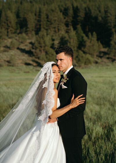 A bride and groom embracing in a grassy field with a forested hillside in the background.