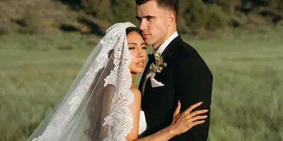 A bride and groom embracing in a grassy field with a forested hillside in the background.