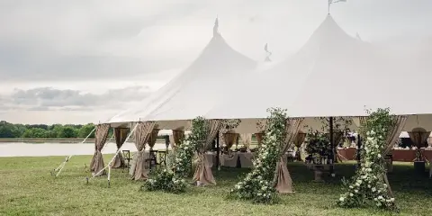 A large white event tent set up on a grassy field with beige draping and floral arrangements.