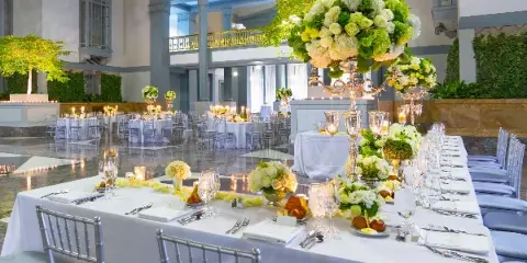 Wedding reception tables set with white linens, silver Chiavari chairs, glassware, flatware, and floral centerpieces in a ballroom.