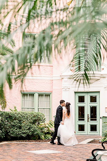 A bride and groom standing together in a courtyard with a pink building in the background.