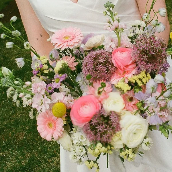 A bride in a white dress holding a large, colorful bouquet of fresh flowers including pink gerberas, ranunculus, and allium.