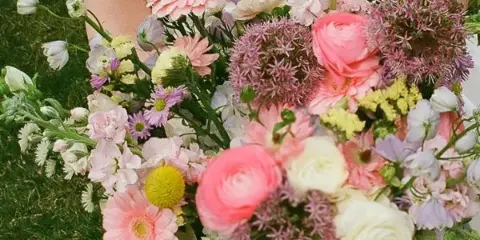A bride in a white dress holding a large, colorful bouquet of fresh flowers including pink gerberas, ranunculus, and allium.