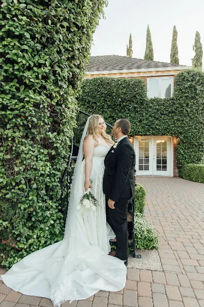 A bride and groom standing together outdoors near a building with ivy-covered walls.