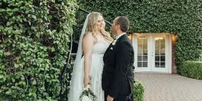 A bride and groom standing together outdoors near a building with ivy-covered walls.
