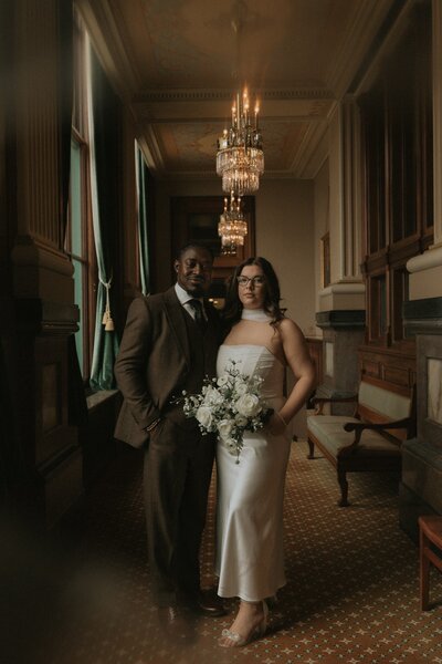 A professional wedding portrait of a couple standing in an elegant hallway with a chandelier.