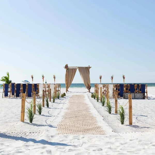 A beach wedding ceremony setup featuring a wooden arch, rows of chairs, and a woven aisle runner on white sand.