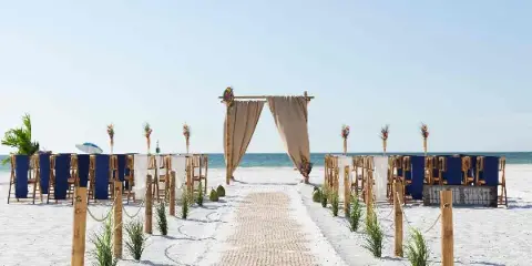 A beach wedding ceremony setup featuring a wooden arch, rows of chairs, and a woven aisle runner on white sand.
