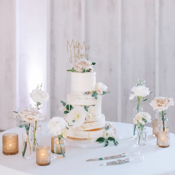 A three-tiered white wedding cake decorated with fresh flowers and a gold 'Mr & Mrs' topper, displayed on a table with candles and floral arrangements.