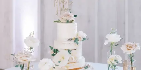 A three-tiered white wedding cake decorated with fresh flowers and a gold 'Mr & Mrs' topper, displayed on a table with candles and floral arrangements.