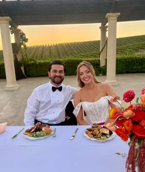 A bride and groom seated at a table with plated food and a floral arrangement at a wedding event.
