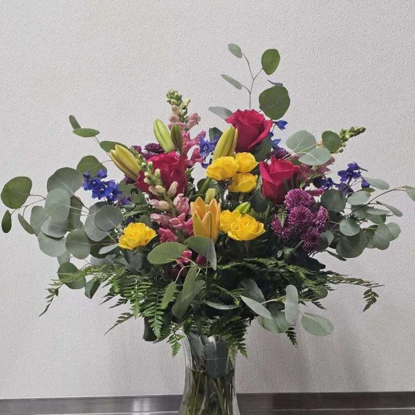 A mixed bouquet of red roses, yellow lilies, and purple flowers with eucalyptus in a clear glass vase.