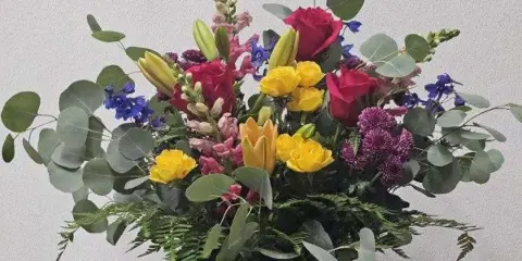 A mixed bouquet of red roses, yellow lilies, and purple flowers with eucalyptus in a clear glass vase.