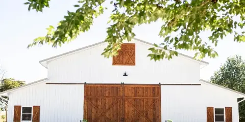 Exterior view of a white barn-style wedding venue with large wooden sliding doors and surrounding greenery.