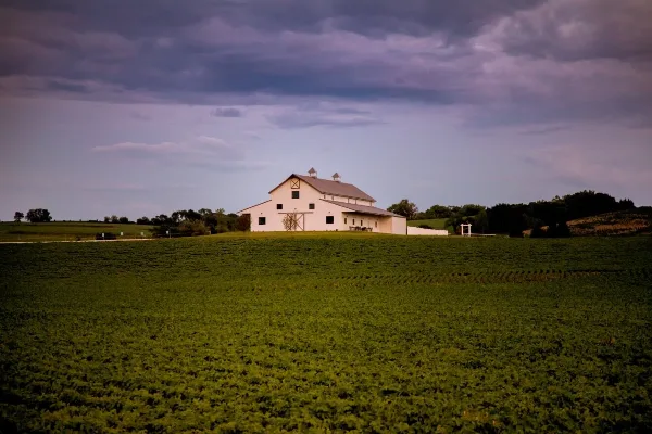 A white barn building with a grey roof situated on a green hill under a cloudy sky.