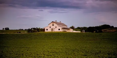A white barn building with a grey roof situated on a green hill under a cloudy sky.