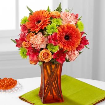 A vibrant floral arrangement featuring orange gerbera daisies, roses, and carnations in an amber glass vase.