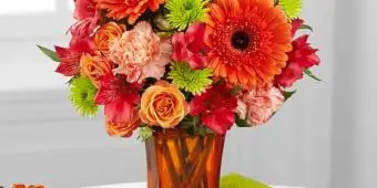 A vibrant floral arrangement featuring orange gerbera daisies, roses, and carnations in an amber glass vase.