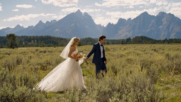 A bride and groom walking through a grassy field with a mountain range in the background.