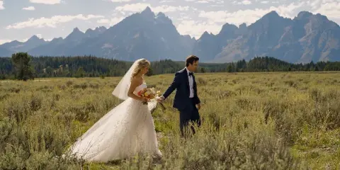 A bride and groom walking through a grassy field with a mountain range in the background.