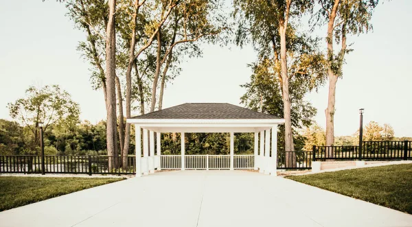 An outdoor white gazebo ceremony site with a paved walkway, surrounded by tall trees and greenery.