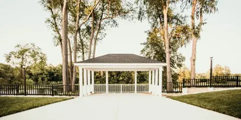 An outdoor white gazebo ceremony site with a paved walkway, surrounded by tall trees and greenery.