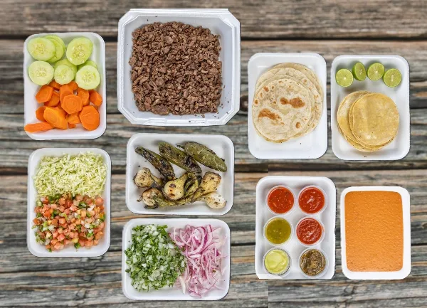 A catering spread featuring carne asada, tortillas, salsas, and garnishes arranged in white takeout containers on a wooden surface.