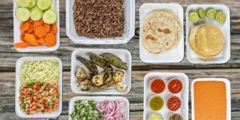A catering spread featuring carne asada, tortillas, salsas, and garnishes arranged in white takeout containers on a wooden surface.