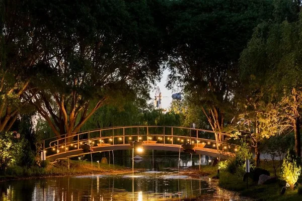 A scenic view of a private lake at dusk, featuring a lit arched bridge, lush greenery, and a windmill in the background.