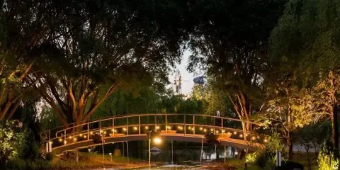 A scenic view of a private lake at dusk, featuring a lit arched bridge, lush greenery, and a windmill in the background.