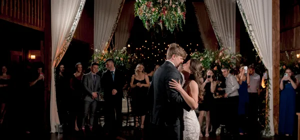 A bride and groom dancing at a wedding reception in a rustic venue with floral decorations and guests watching.