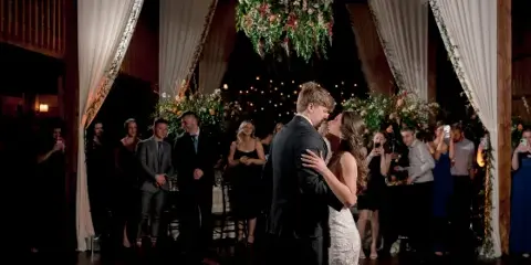 A bride and groom dancing at a wedding reception in a rustic venue with floral decorations and guests watching.