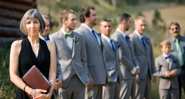 A woman in a black dress holding a book stands in front of a groom and groomsmen in grey suits at an outdoor wedding.